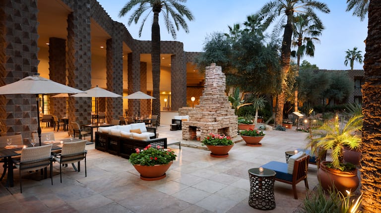 Tables With Umbrellas, Soft Seating, Furnace, and Candlelit Tables on Outdoor Patio Surrounded by Palm Trees at Dusk