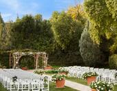 Daytime View of White Chairs Arranged Theater Style, White Aisle Runner, Large Flower Pots, and Archway Set Up For Garden Wedding