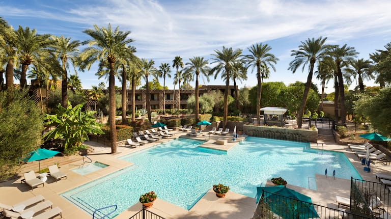 Vista de día de sombrillas verdes, sillas reclinables junto a la piscina norte al aire libre rodeada de palmeras y fachada del hotel en el fondo
