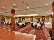 Dance Floor Surrounded by Banquet Tables With Place Settings, White Flowers, White Napkins, Drinking Glasses, and White Linens, Black Chairs, Food Service Area. and Cake Table Set Up in Ballroom For Wedding