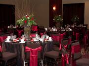Red Roses, Red Candles, Place Settings, and Drinking Glasses on Dining Tables With Decorative Linens and Chairs With Red Sashes Set Up in Ballroom For Wedding