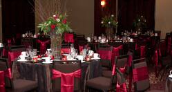 Red Roses, Red Candles, Place Settings, and Drinking Glasses on Dining Tables With Decorative Linens and Chairs With Red Sashes Set Up in Ballroom For Wedding