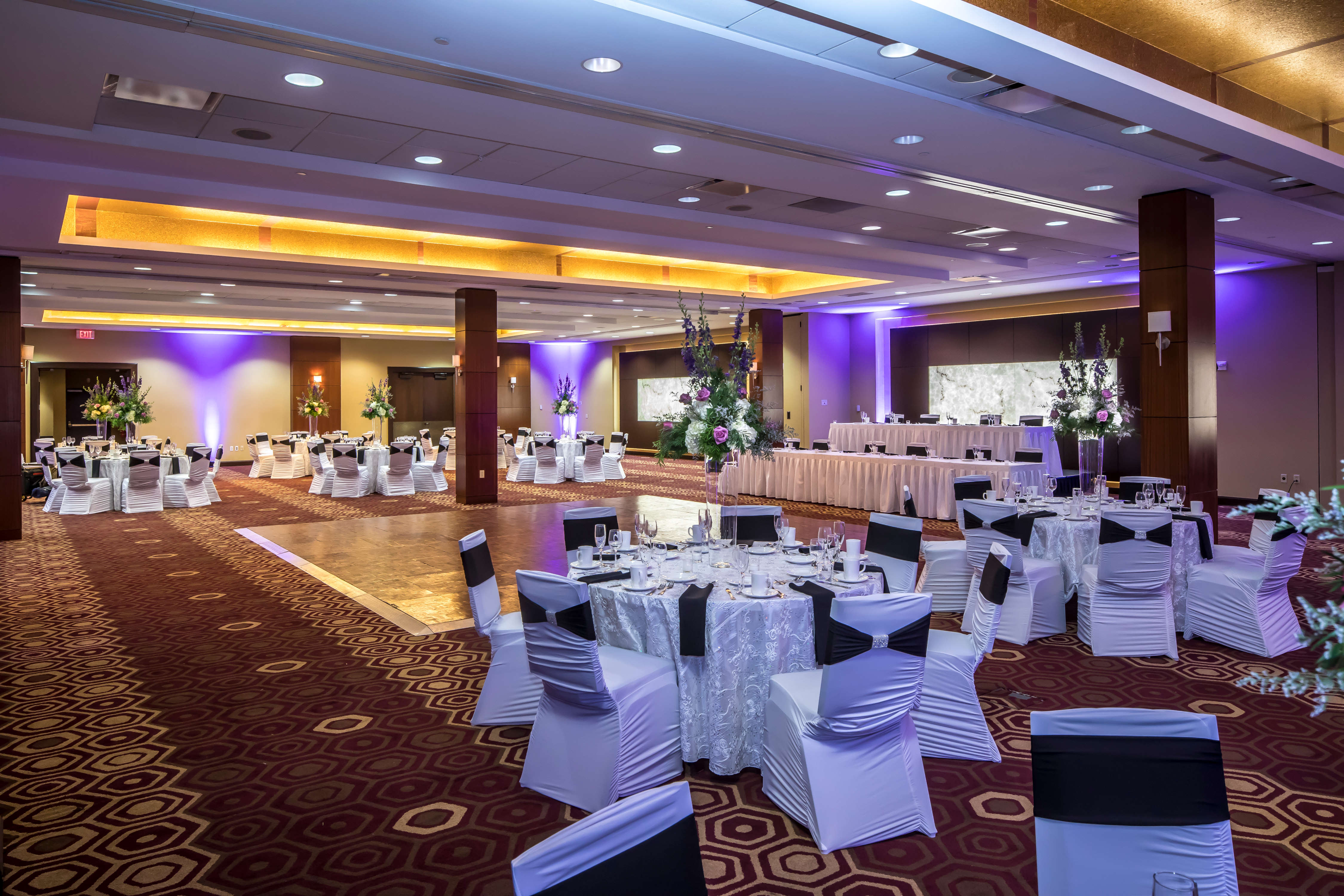 Dance Floor Surrounded by Banquet Tables With Place Settings, Flowers, Drinking Glasses, and White Linens, White Chairs With Black Sashes, Two Head Tables, and Purple Lighting in Ballroom Set Up For Wedding