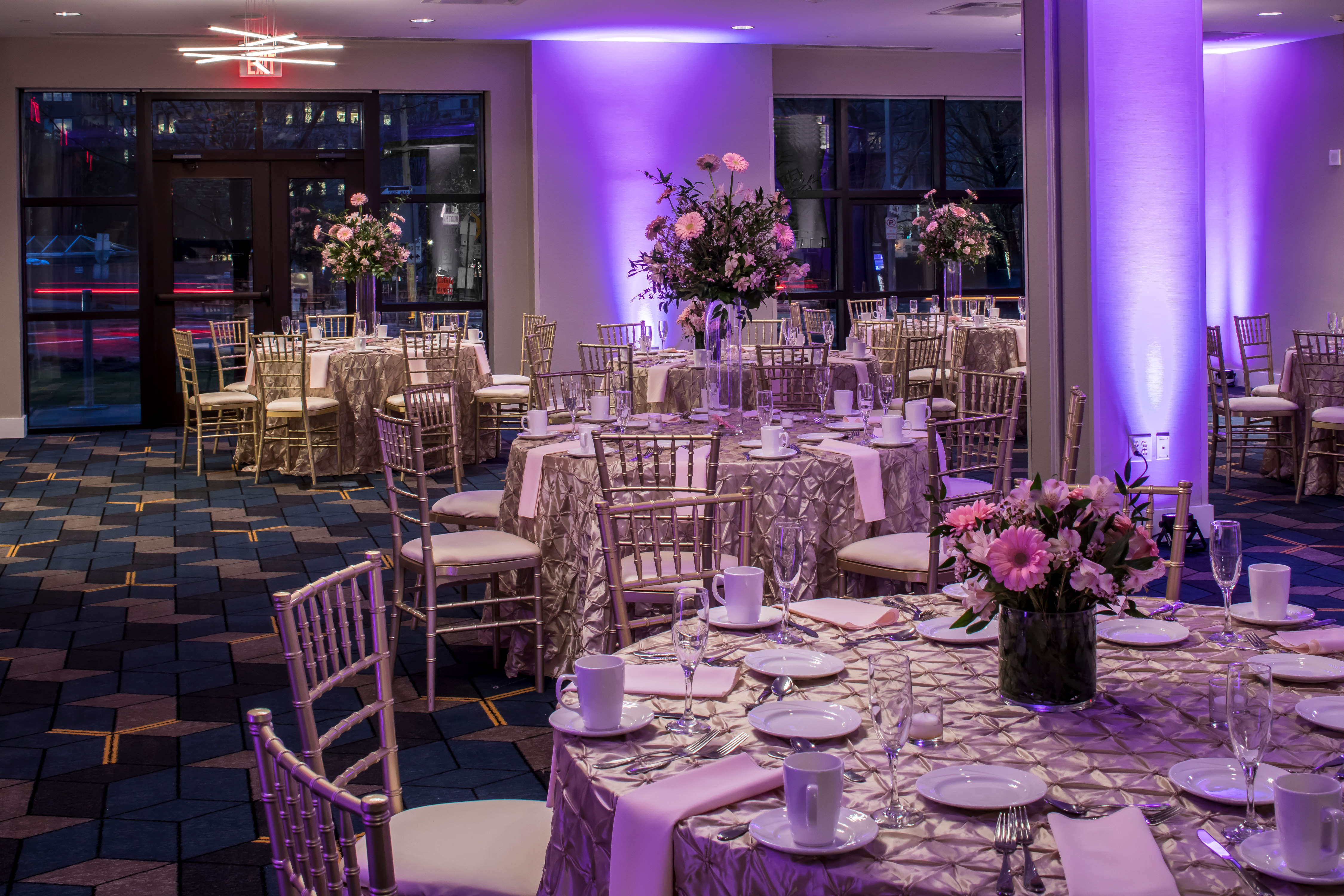 Ballroom With Purple Lighting, Place Settings, Flowers, Drinking Glasses, White Napkins, and Decorative Linens on Round Tables, Chairs, Windows With Sheer Drapes, and Glass Entry Doors