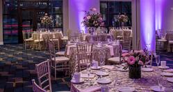 Ballroom With Purple Lighting, Place Settings, Flowers, Drinking Glasses, White Napkins, and Decorative Linens on Round Tables, Chairs, Windows With Sheer Drapes, and Glass Entry Doors