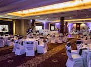 Banquet Tables With Place Settings, Flowers, Drinking Glasses, and White Linens, White Chairs With Black Sashes, Dance Floor, Two Head Tables, Three Presentation Screens, and Purple Lighting in Ballroom Set Up For Wedding