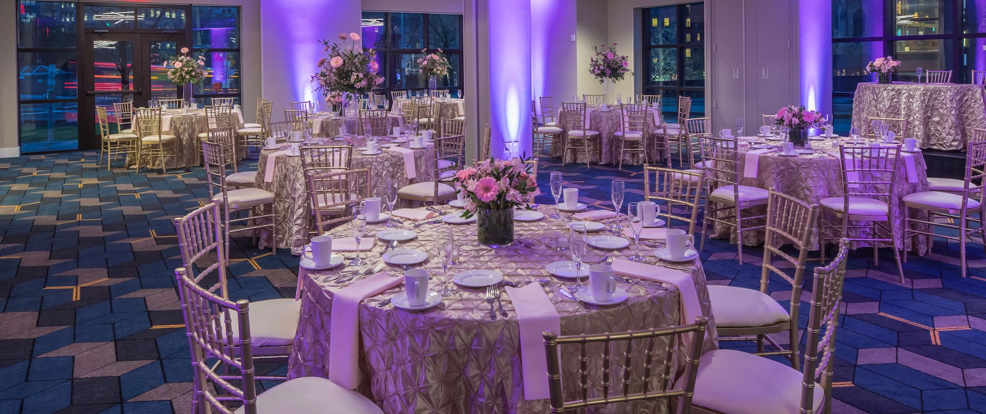 Ballroom With Purple Lighting, Place Settings, Flowers, Drinking Glasses, White Napkins, and Decorative Linens on Round Tables, Chairs, Windows and Glass Entry Doors