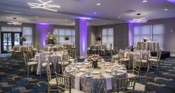 Ballroom With Purple Lighting, Place Settings, Flowers, Drinking Glasses, White Napkins, and Decorative Linens on Round Tables, Chairs, Windows With Sheer Drapes, and Entry Doors