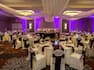 Banquet Tables With Place Settings, Candles, Flowers, Drinking Glasses, Burgundy Napkins, and White Linens, White Chairs With Burgundy Sashes, Buffet Table on Dance Floor, Head Table, and Purple Lighting in Ballroom Set Up For Wedding