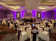 Banquet Tables With Place Settings, Candles, Flowers, Drinking Glasses, Burgundy Napkins, and White Linens, White Chairs With Burgundy Sashes, Buffet Table on Dance Floor, Head Table, and Purple Lighting in Ballroom Set Up For Wedding