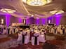 Banquet Tables With Place Settings, Candles, Flowers, Drinking Glasses, Burgundy Napkins, and White Linens, White Chairs With Burgundy Sashes, Buffet Table on Dance Floor, Head Table, Draped Ceiling, and Purple Lighting in Ballroom Set Up For Wedding