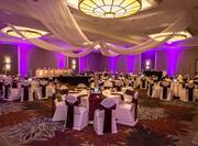Banquet Tables With Place Settings, Candles, Flowers, Drinking Glasses, Burgundy Napkins, and White Linens, White Chairs With Burgundy Sashes, Buffet Table on Dance Floor, Head Table, Draped Ceiling, and Purple Lighting in Ballroom Set Up For Wedding