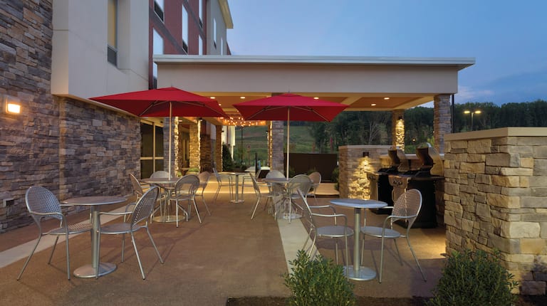 Illuminated Outdoor Patio With Two Grills and Chairs Around Round Table With Red Umbrellas at Dusk