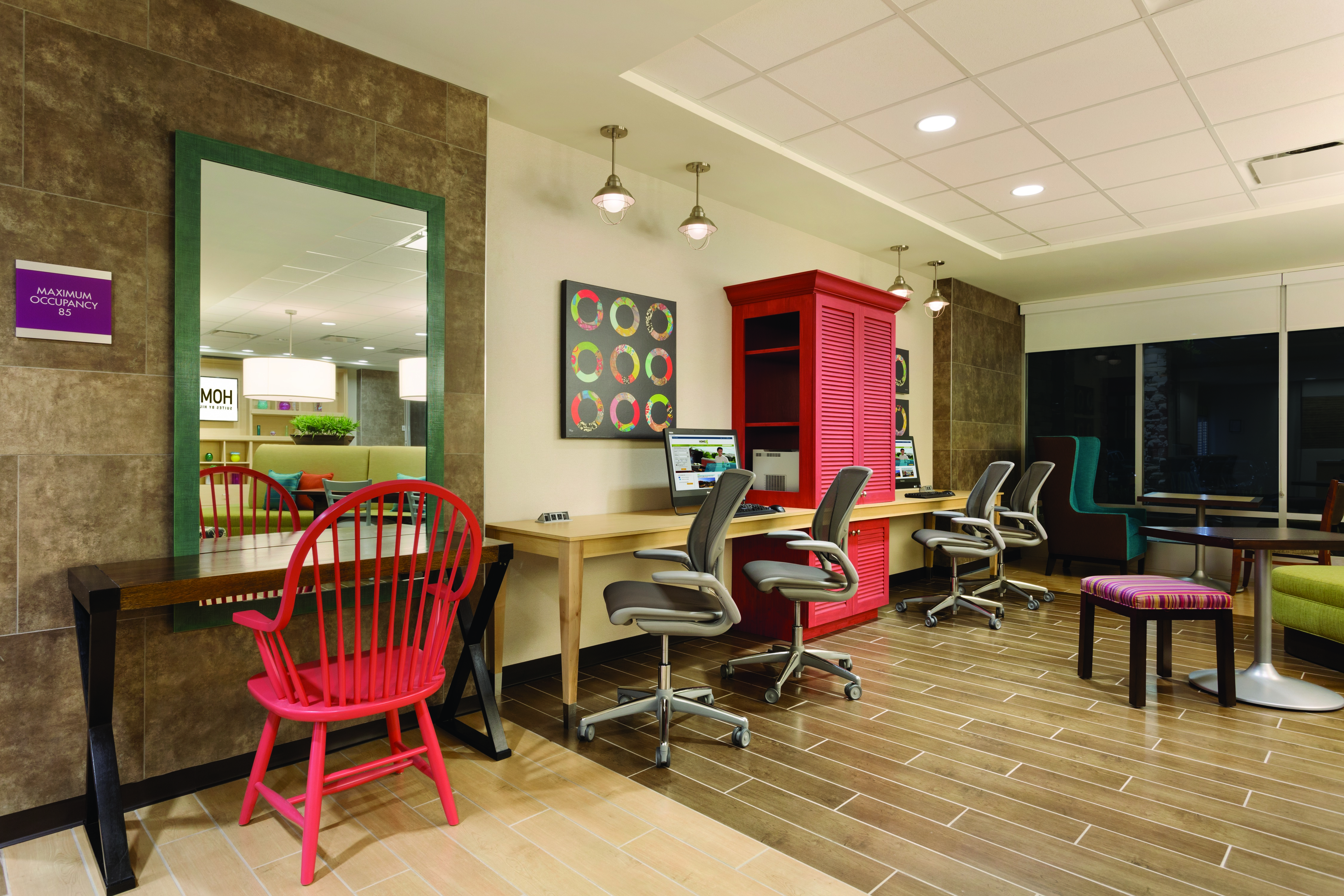 Occupancy Sign by Large Mirror Above Small Desk With Red Wooden Chair, Colorful Wall Art, Red Storage Cabinet Between Two Computers on Long Desk With Four Ergonomic Chairs and Additional Seating by Window in Business Center