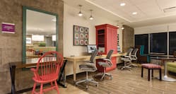 Occupancy Sign by Large Mirror Above Small Desk With Red Wooden Chair, Colorful Wall Art, Red Storage Cabinet Between Two Computers on Long Desk With Four Ergonomic Chairs and Additional Seating by Window in Business Center