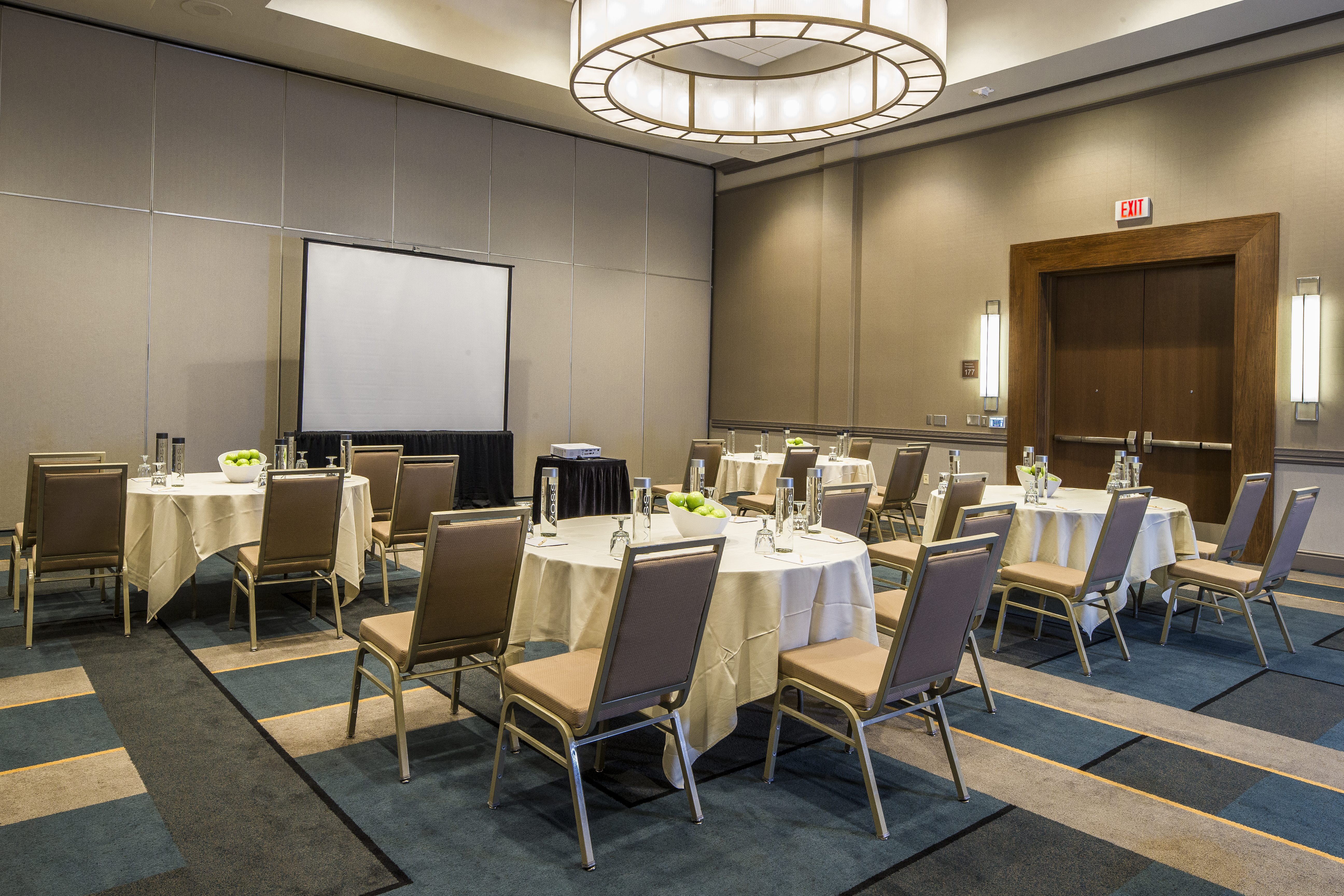 Meeting Room With Water Bottles, Bowl of Green Apples, Drinking Glasses, Notepads and Linens on Banquet Tables, Chairs, Projector Table, Presentation Screen, and Double Entry Doors