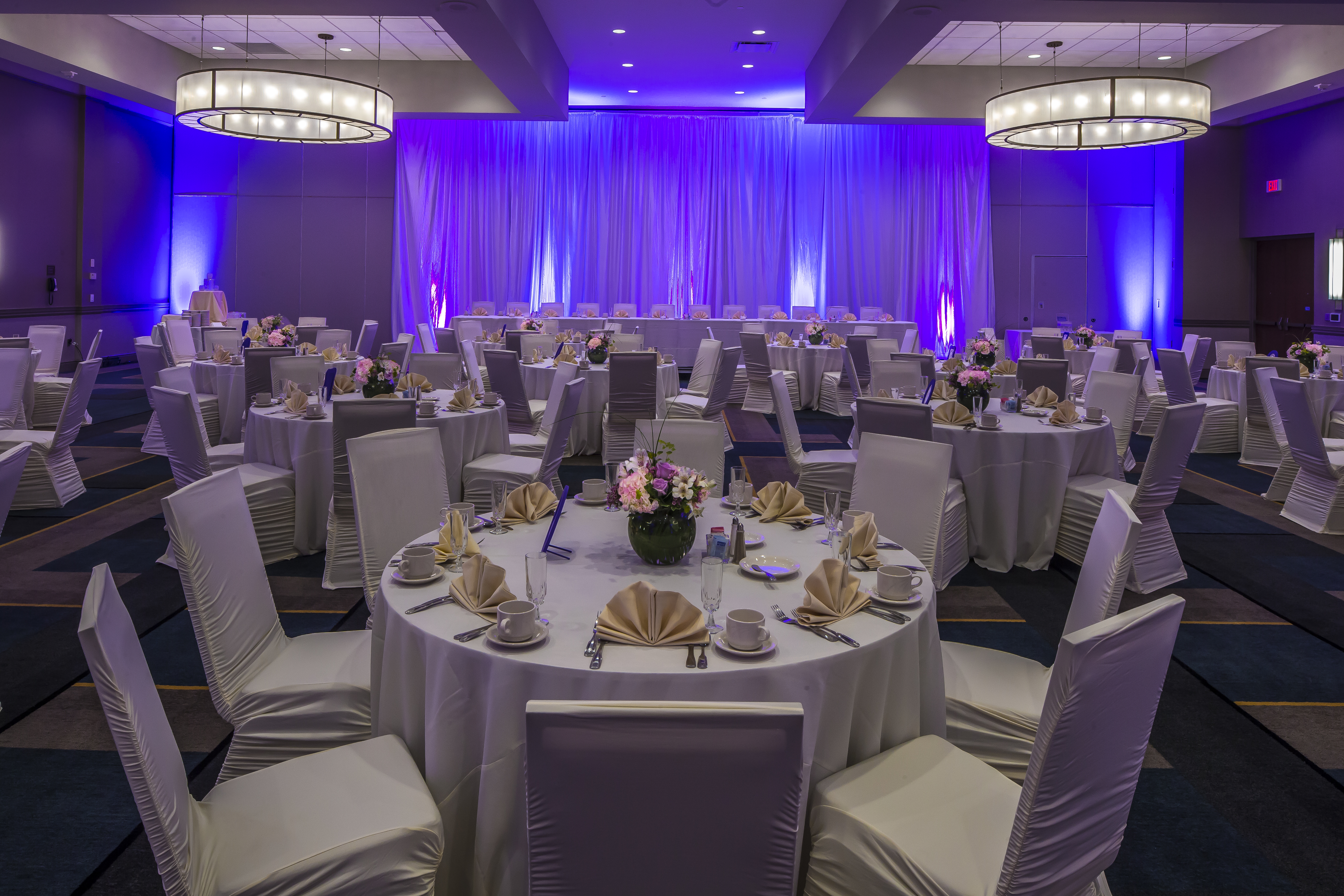 White Chairs, Place Settings, Flowers, Fan-Folded Napkins, Drinking Glasses, and White Linens on Round Tables, and Dramatic Drapes With Purple Lighting Behind Head Table in Banquet Hall Set Up For Wedding Reception