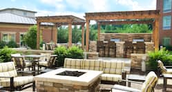 Outdoor patio with fire pit, surrounded by lounge chairs and sofa, with BBQ grills under pavilion in background