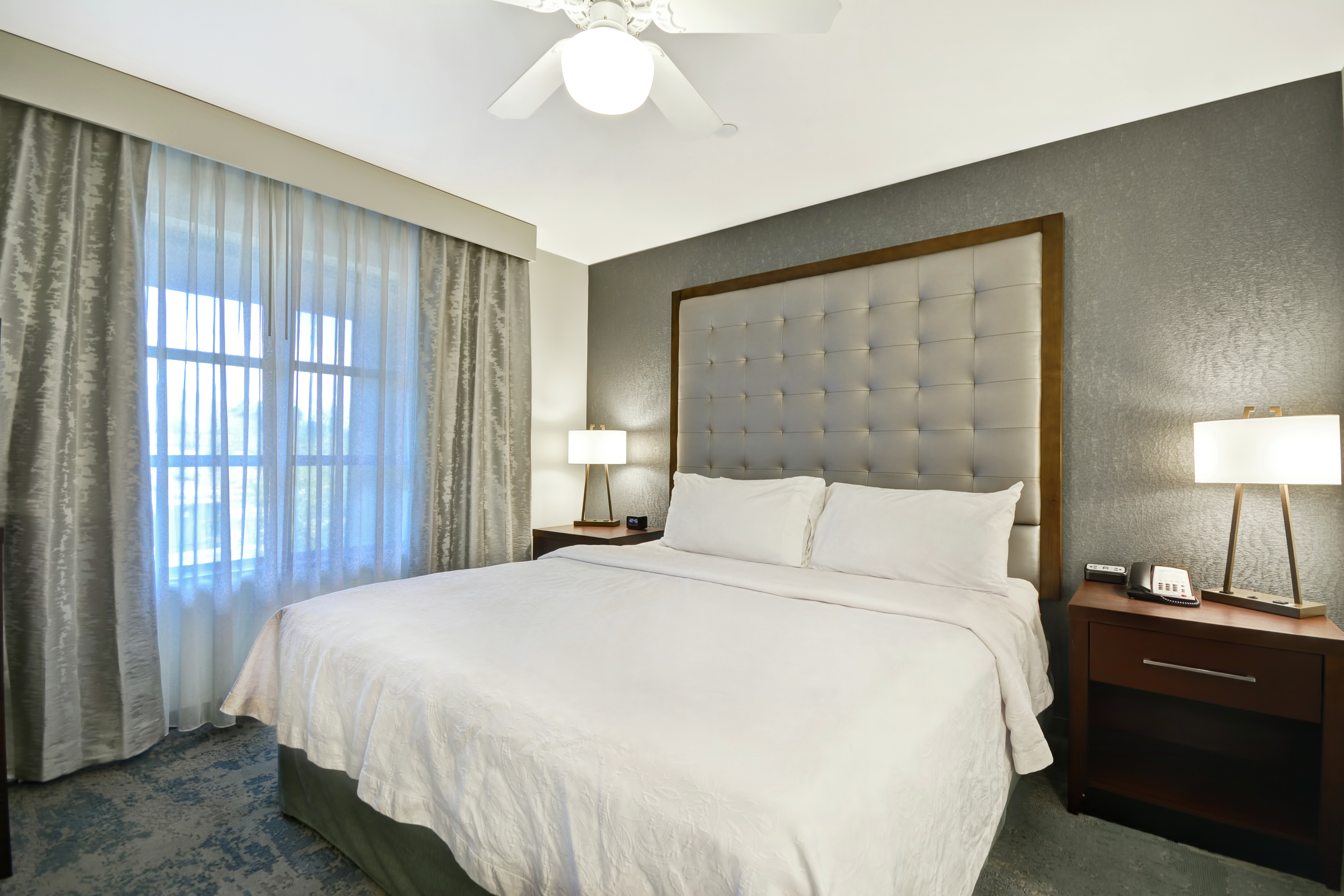 King Bed Between Two Illuminated Lamps by Large Window With Long Drapes and Ceiling Fan in Studio Suite