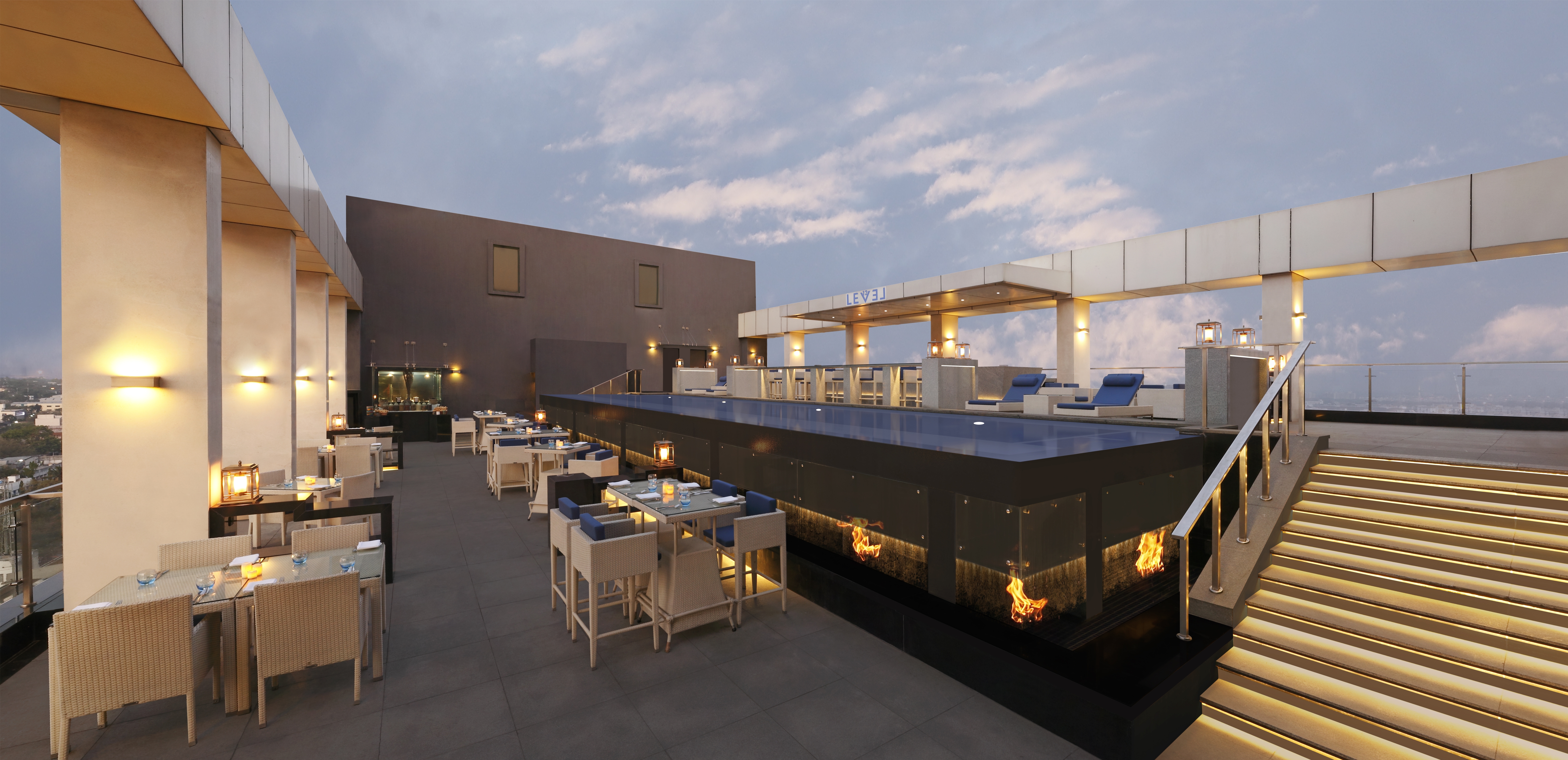 Loungers by Rooftop Swimming Pool and Staircase Down to Candlelit Tables and Chairs by Large Windows in Lounge Area