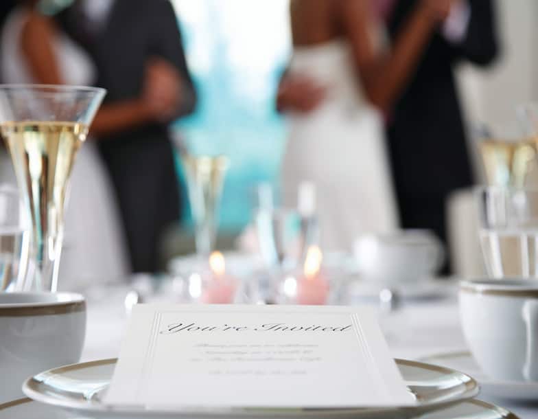 Detailed View of Wedding Invitation, Place Settings, and Champagne Flutes on Table With White Linens, and View of People Dancing in Background