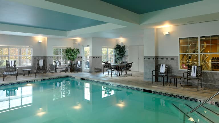Indoor Pool with lounge chairs and dining table, and partial view of fitness center through indoor window