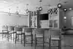 Bar counter lined with green barstools beneath globe pendant lights, with bottle cabinets and a TV behind the bar.