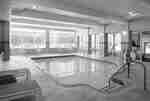 Indoor swimming pool surrounded by a tiled deck, lounge chairs, small tables, and large windows.