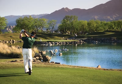 A golfer taking a shot over a lake with mountains in the backgrounds