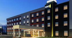 Modern Home2 Suites hotel exterior featuring covered patio, glowing guest room windows, and beautiful dusk sky.