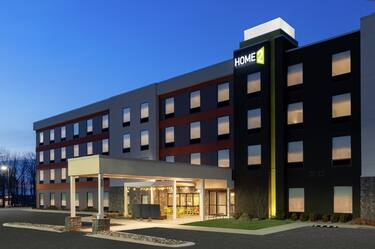Modern Home2 Suites hotel exterior featuring covered patio, glowing guest room windows, and beautiful dusk sky.