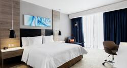 Wall Art and King Bed Between Two Illuminated Lamps and Bedside Tables, Reading Chair, Illuminated Floor Lamp, Window With Long Drapes, and Work Desk in Guest Room