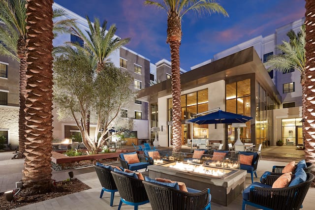 Evening View of the Illuminated Outdoor Patio Fire Pit Surrounded by Palm trees, and Seating for Ten with Orange Pillows and Two Patio Umbrellas in the Background