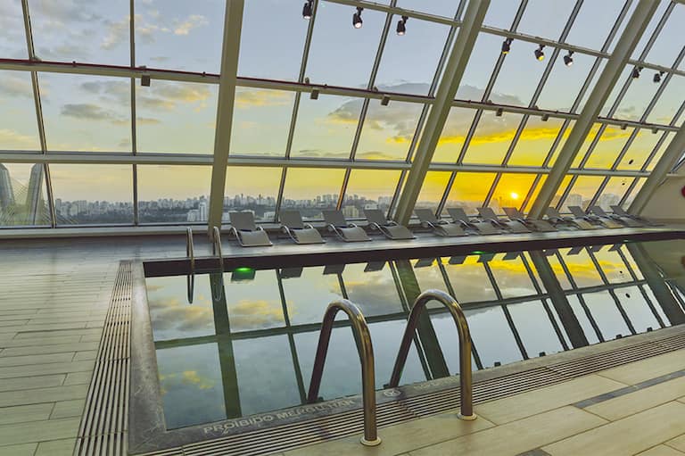 Área da piscina coberta com espreguiçadeiras e janelas amplas com vista para o pôr do sol