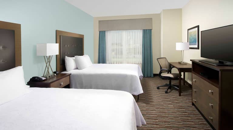 Guestroom With Ceiling Fan Above Two Queen Beds, Window With Long Drapes, Work Desk and TV