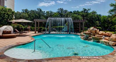 Daytime View of Outdoor Pool Facing Waterfall With Lounge Seating, Tables and Sun Umbrella Surrounded by Trees