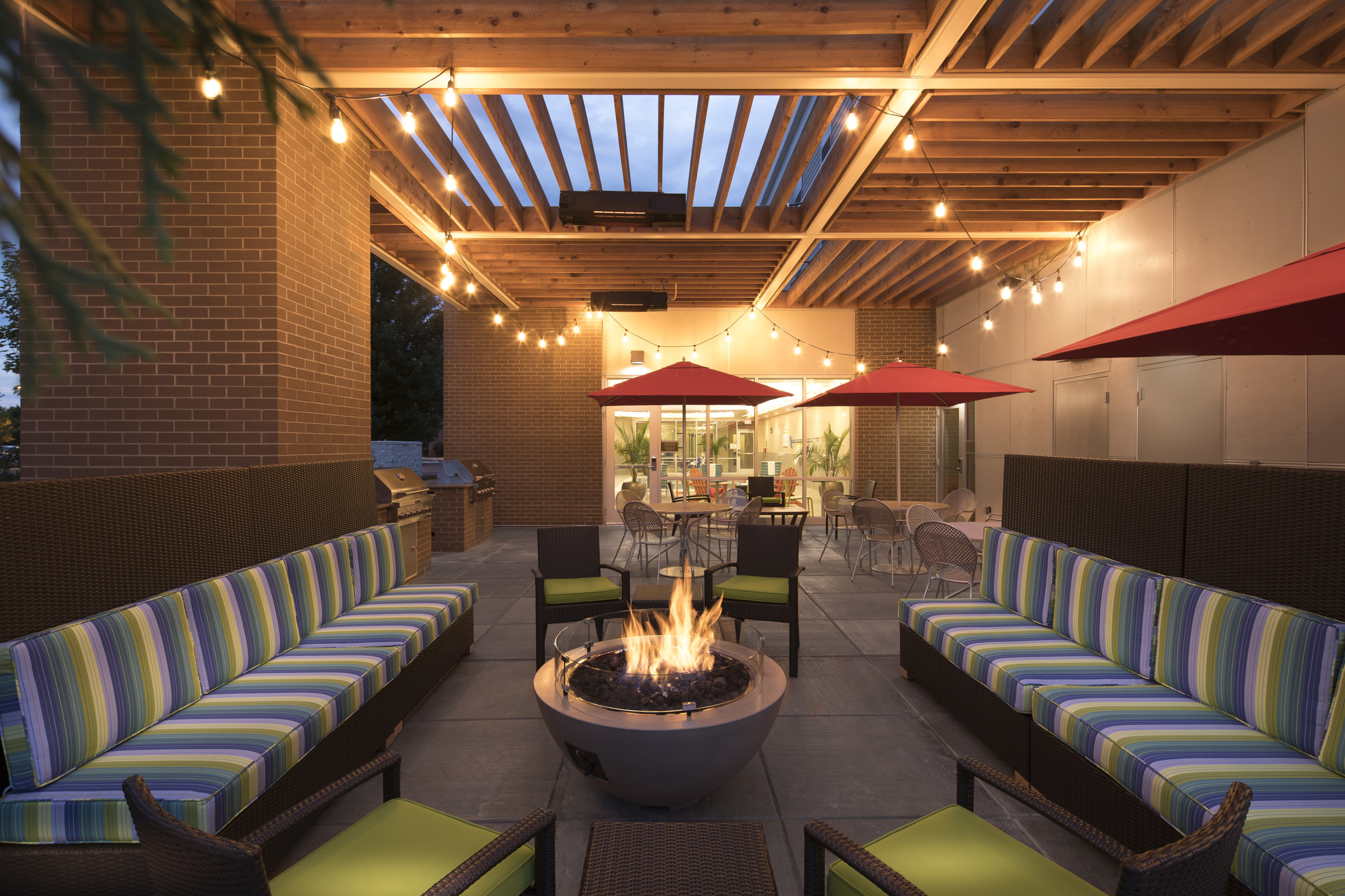 Outdoor patio area lit up with sofas and lounge chairs surrounding fire pit, and tables with umbrella covers
