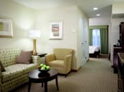 Bowl of Apples on Coffee Table, Wall Art Above Sofa, Illuminated Lamp on Side Table, Armchair, Open Doorway to View of Bedroom, Hospitality Center, and TV in Suite Living Area
