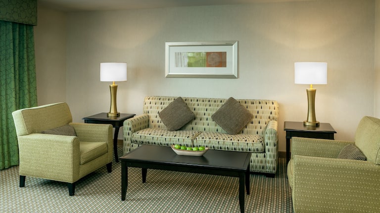Window With Green Drapes, Two Armchairs, Illuminated Lamps on Side Tables, and Wall Art Above Sofa in  Suite Living Room