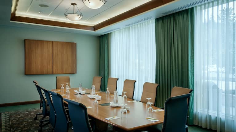 Seating for 12, Water Pitchers, Drinking Glasses, and Notepads at Connectivity Table in Boardroom With Media Cabinet, and Windows With Long Drapes