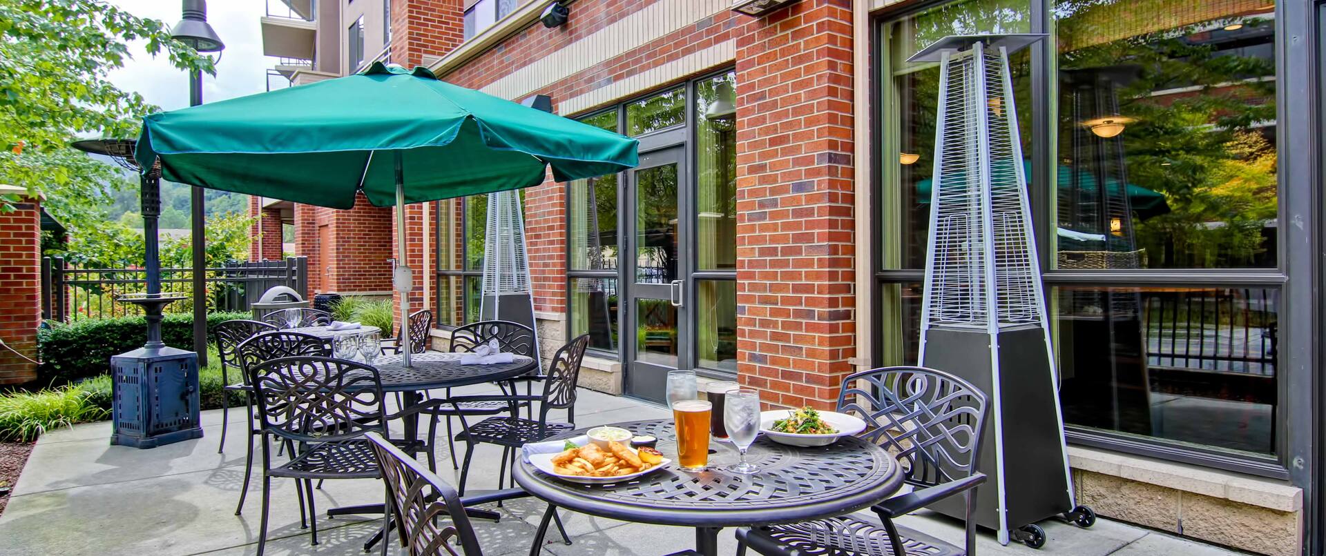 Green Umbrella, Table With Plates of Food and Drinks, Chairs, and Heat Lanterns on Patio by Hotel Exterior