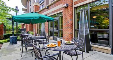 Green Umbrella, Table With Plates of Food and Drinks, Chairs, and Heat Lanterns on Patio by Hotel Exterior