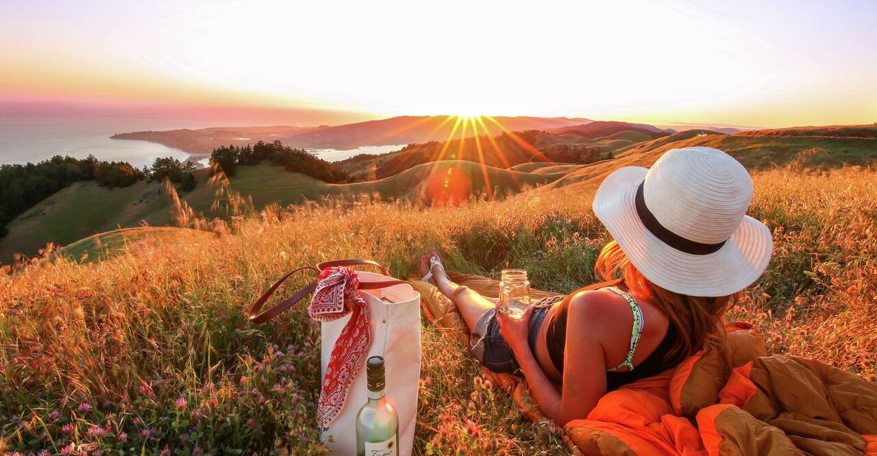  Woman lying in a meadow drinking wine while watching the sun set over the ocean.