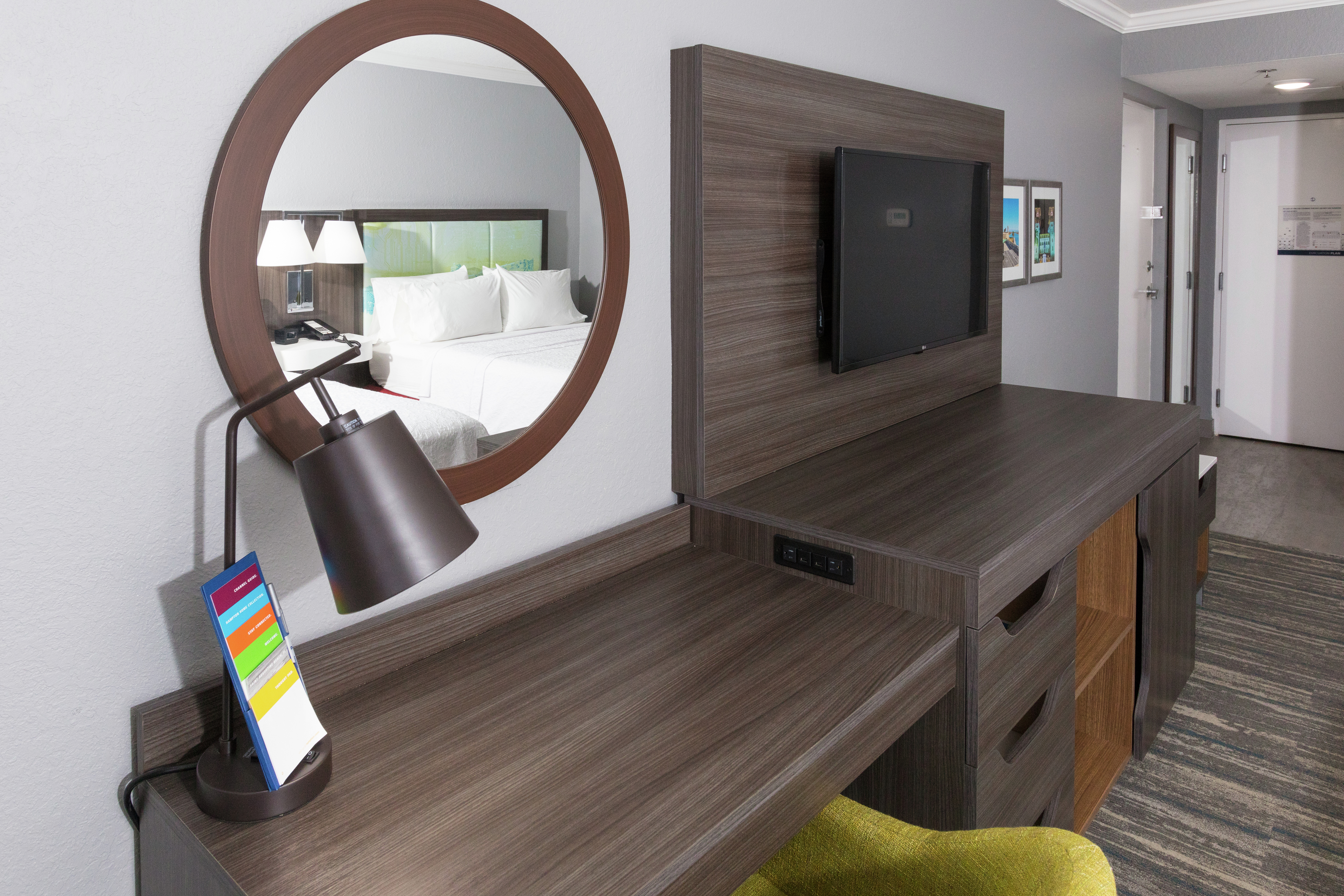 Bed Reflected in Round Wall Mirror Above Work Desk Next to Flat Screen TV in Guest Room