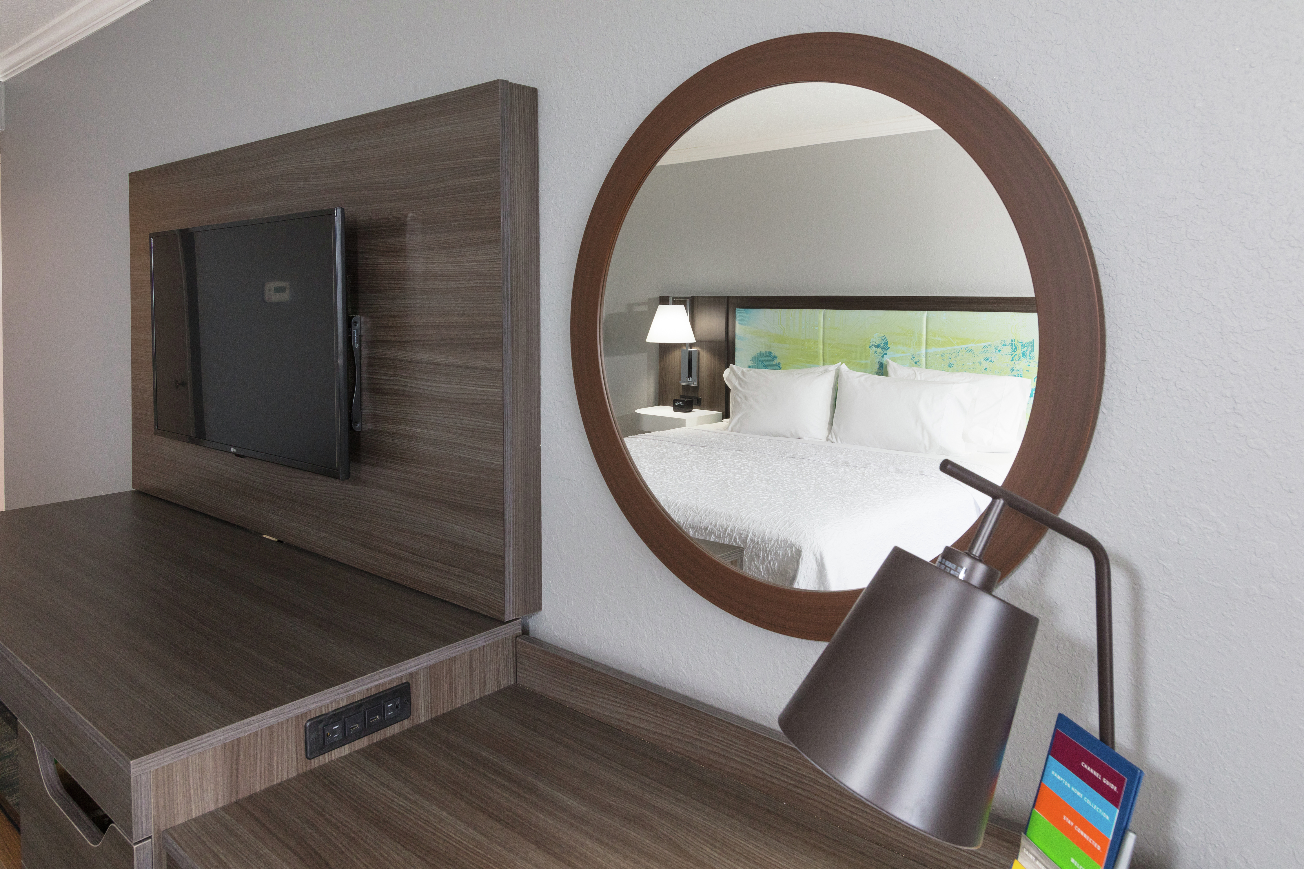 King Bed Reflected in Round Wall Mirror Above Work Desk in Guest Room with Wall-Mounted Flat Screen TV