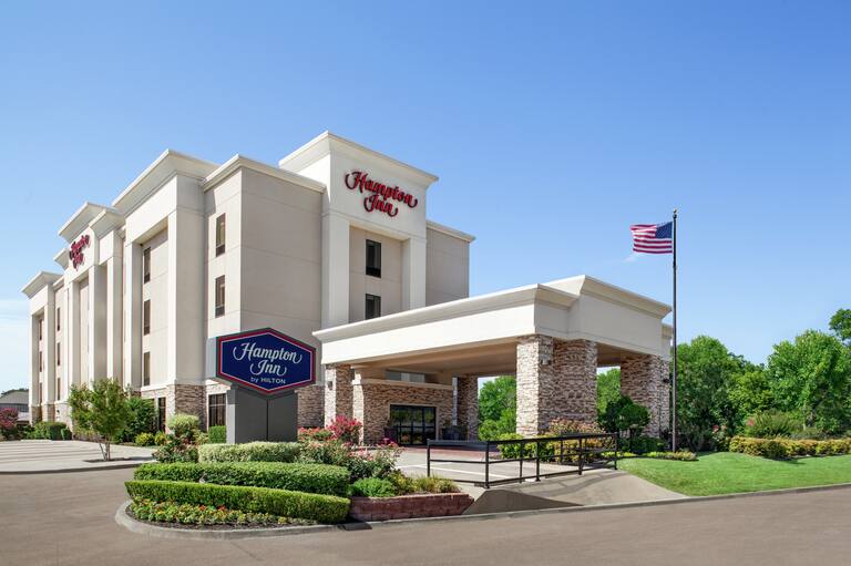 Welcoming Hampton Inn hotel exterior featuring lush greenery, porte cochere, and blue sky.