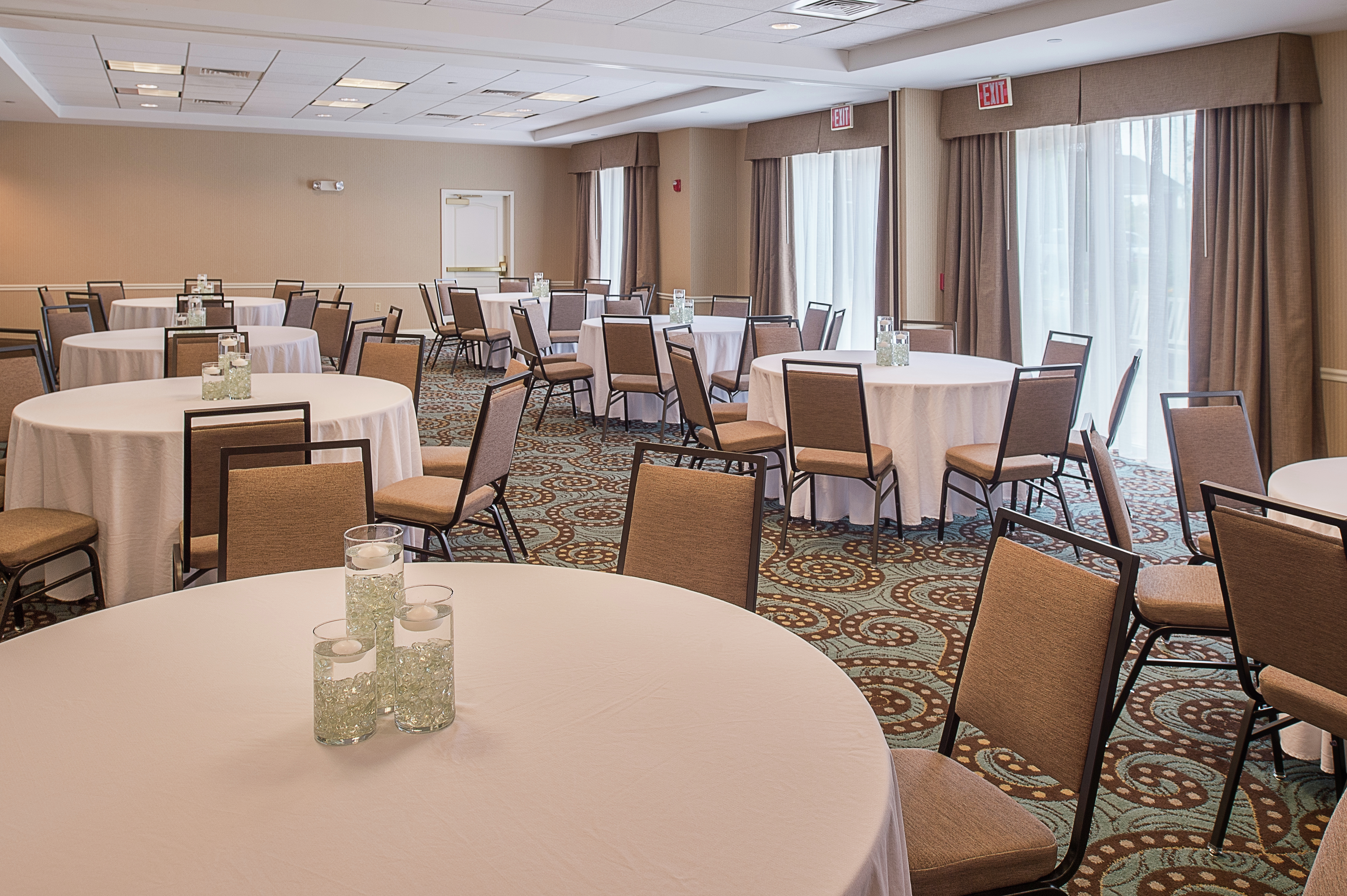Tan Chairs at Round Tables With Candles on White Linens and Windows With Long Drapes in Meeting Space