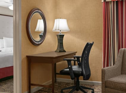 Suite living area featuring work desk with ergonomic chair, mirror, and private bedroom with two comfortable queen beds in background.
