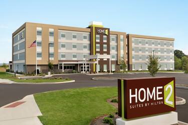 Modern Home2 Suites hotel exterior featuring entry sign, large parking lot, and bright blue skies.