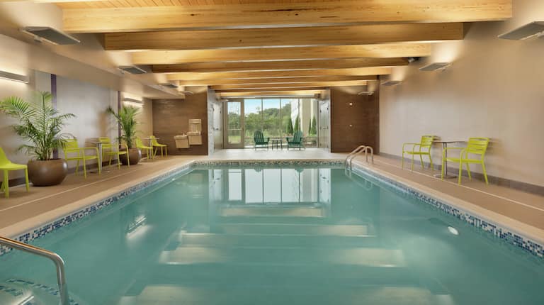Large indoor swimming pool featuring beautiful outside view, patio chairs, and cedar wooden beams.